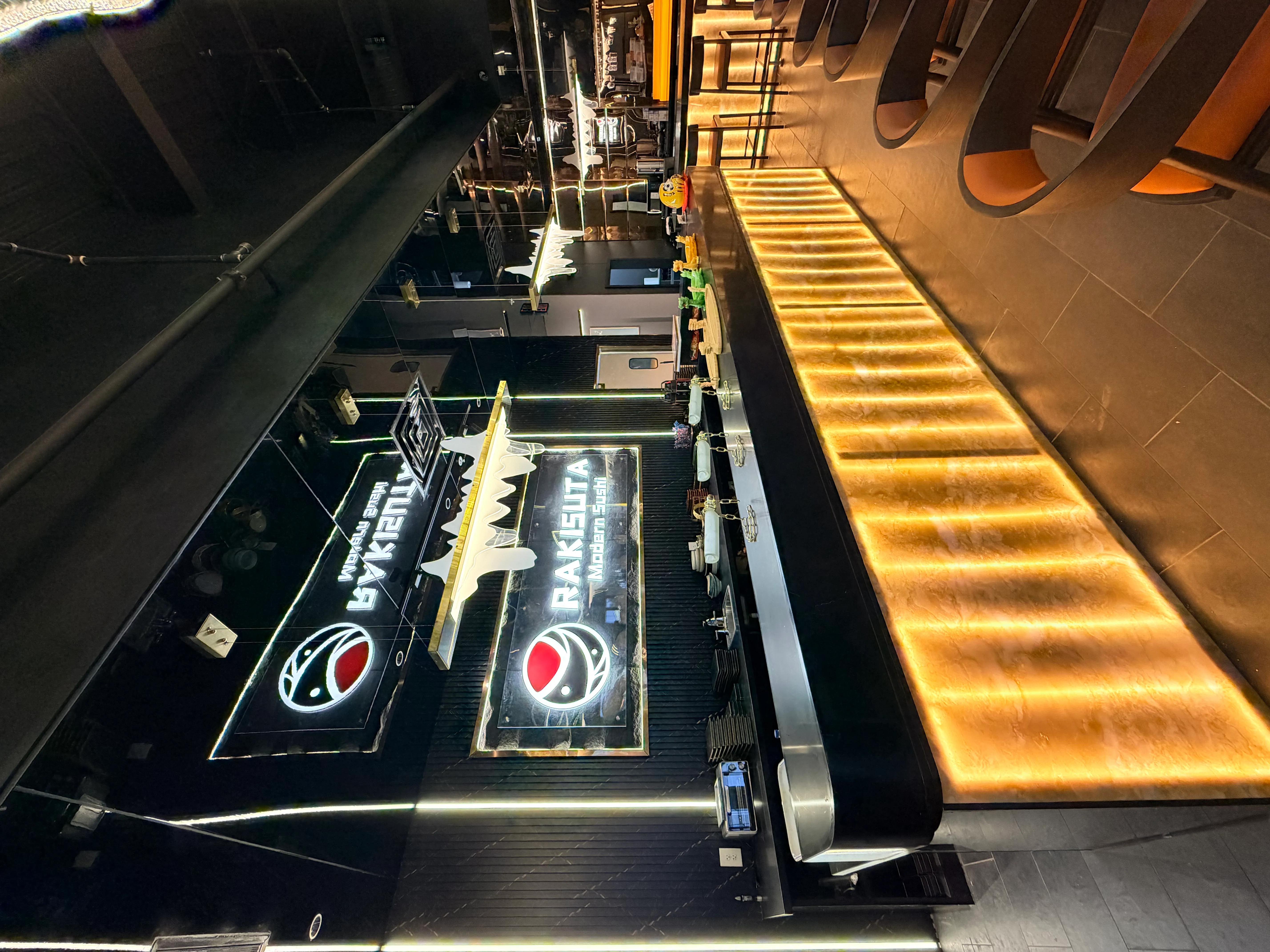A closer view of the Rakisuta bar area, highlighting the illuminated logo, counter setup, and decorative elements like Daruma dolls and chain-linked sushi displays.