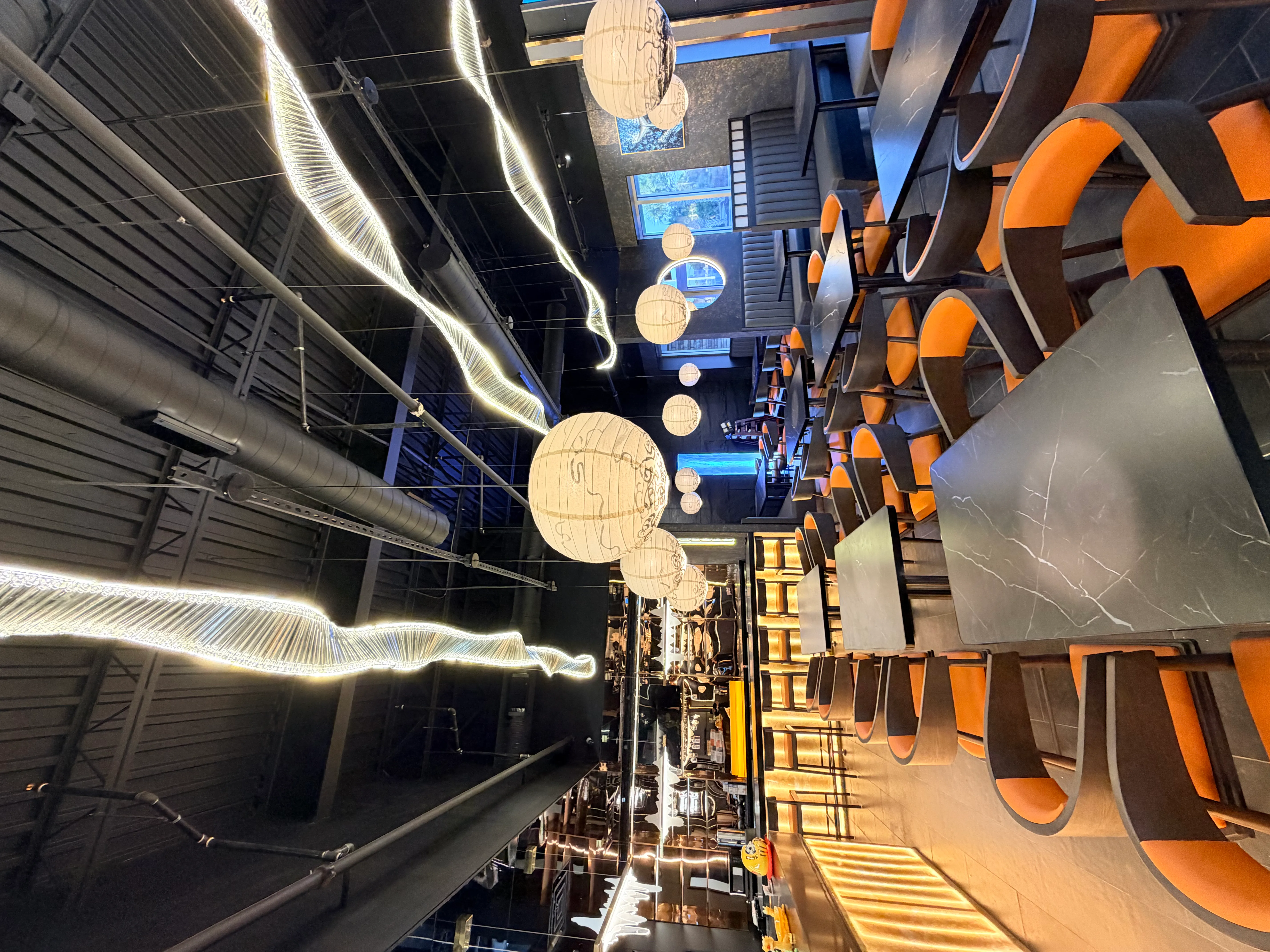 The dining room of Rakisuta, showcasing modern black marble tables, orange-upholstered chairs, hanging paper lanterns, and dramatic LED light fixtures against an industrial-style ceiling.